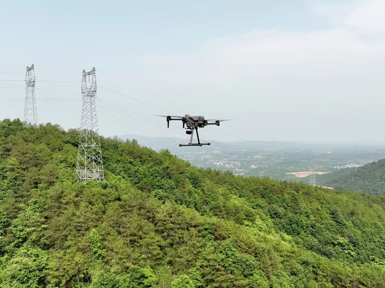 An S400E drone flying beside power-line pylons on a forested ridge